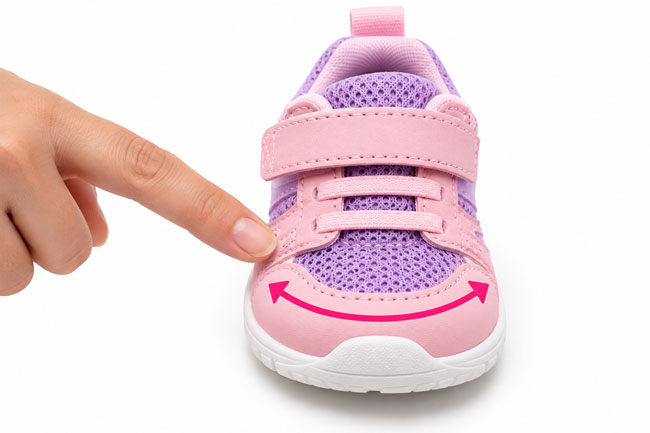 Finger pointing to the wide, rounded toe box of a pink and purple baby shoe, highlighting extra room for natural toe movement and comfortable fit.