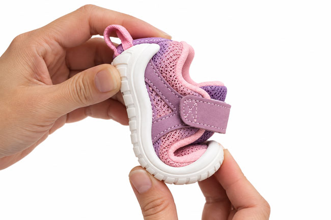 Hands bending a pink and purple baby shoe to demonstrate its flexible sole and lightweight construction for babies learning to walk.